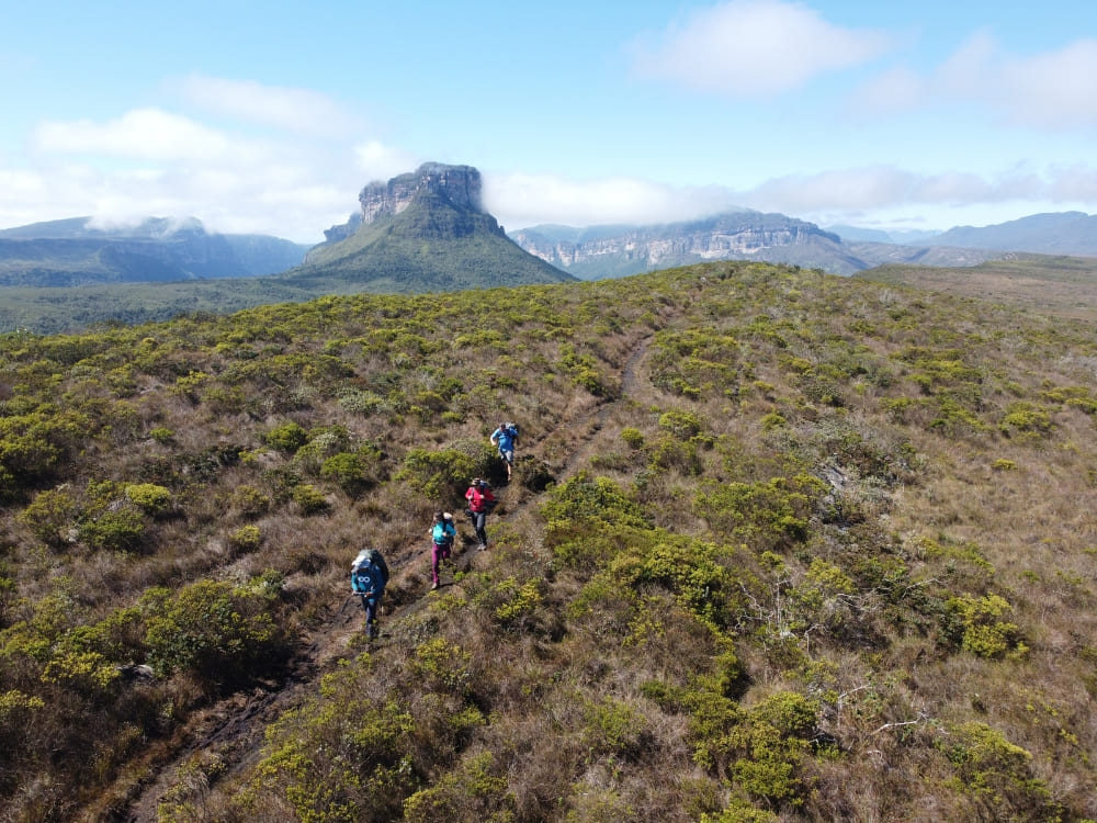 A group of hikers trekking on a dirt trail across a green landscape towards a flat-topped mountain in Vale do Pati. A group of hikers trekking on a dirt trail across a green landscape towards a flat-topped mountain in Vale do Pati.