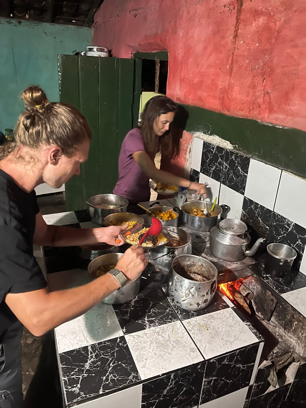Two hikers serving themselves fresh homemade food from various pots in a rustic homestay kitchen. Two hikers serving themselves fresh homemade food from various pots in a rustic homestay kitchen.