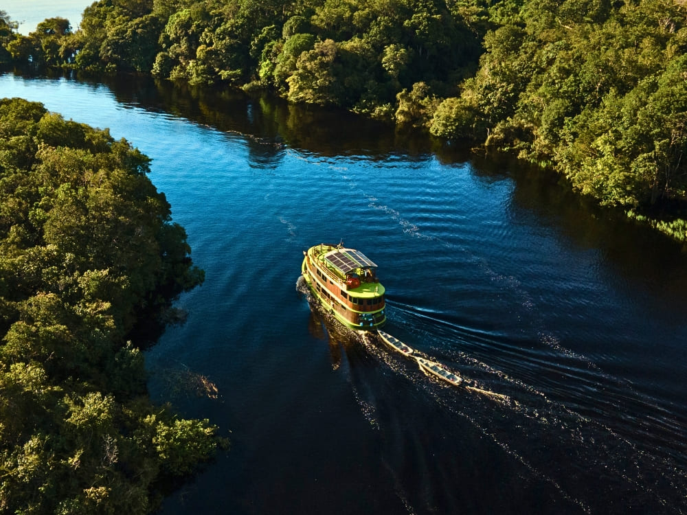 A green ecotourism riverboat with solar panels cruises down a dark river surrounded by lush Amazon rainforest. A green ecotourism riverboat with solar panels cruises down a dark river surrounded by lush Amazon rainforest.