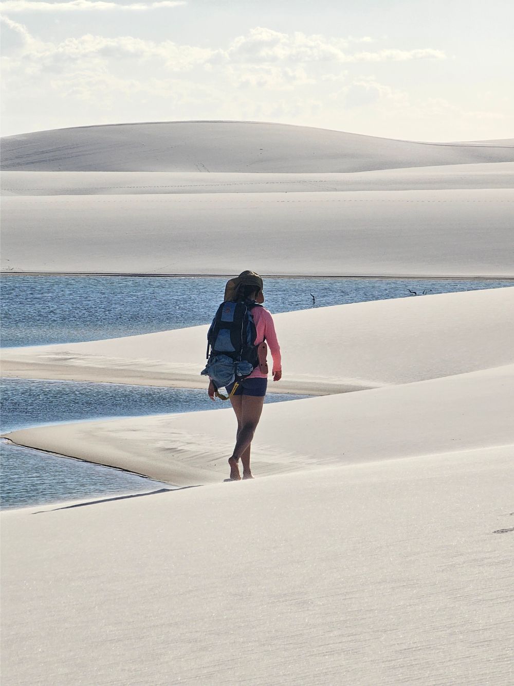 A backpacker walks barefoot along the white sand dunes near a blue rainwater lagoon while trekking in Lençóis Maranhenses National Park. A backpacker walks barefoot along the white sand dunes near a blue rainwater lagoon while trekking in Lençóis Maranhenses National Park.