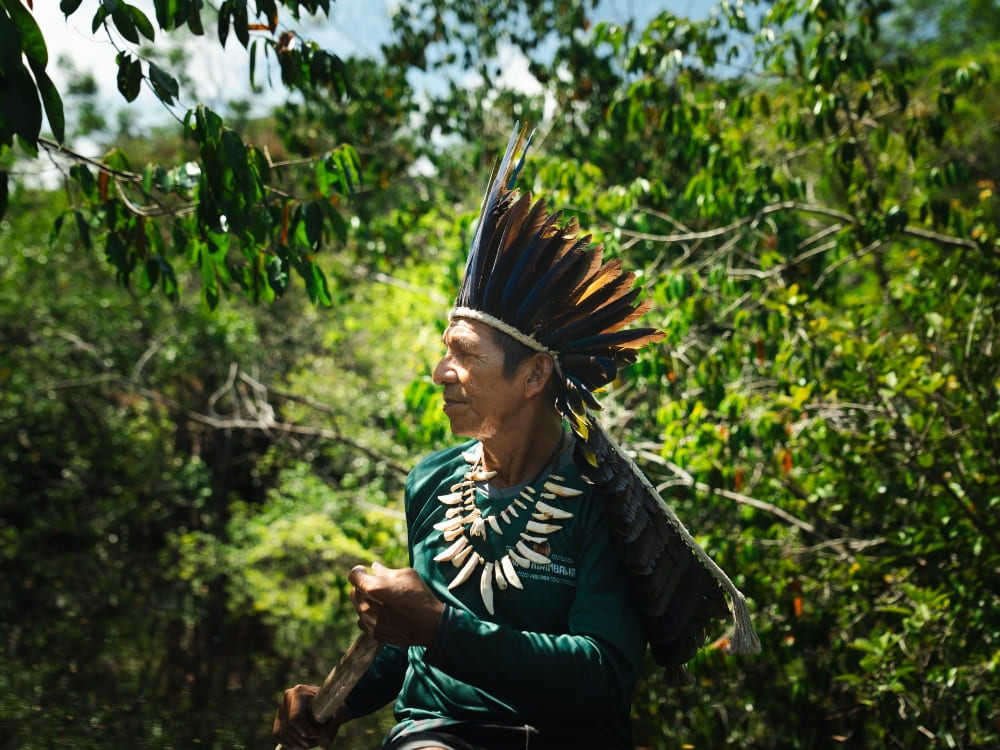 An Indigenous man wearing a traditional feather headdress walks through the lush green Amazon jungle. An Indigenous man wearing a traditional feather headdress walks through the lush green Amazon jungle.