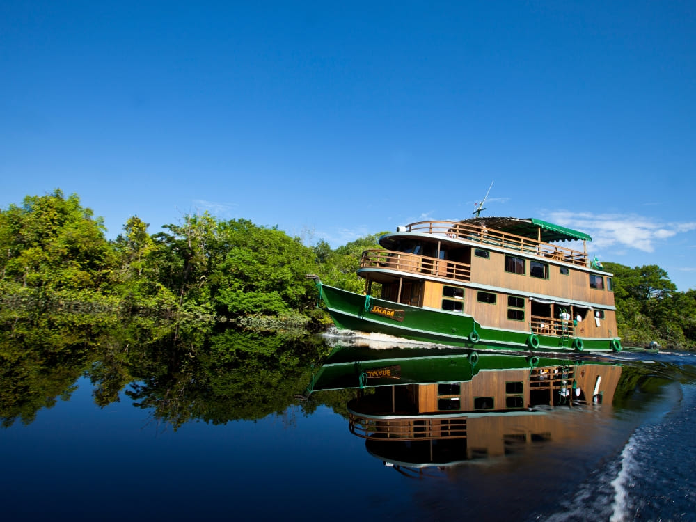 A rustic, three-deck wooden riverboat cruises smoothly along a dark Amazonian river beneath a blue sky. A rustic, three-deck wooden riverboat cruises smoothly along a dark Amazonian river beneath a blue sky.