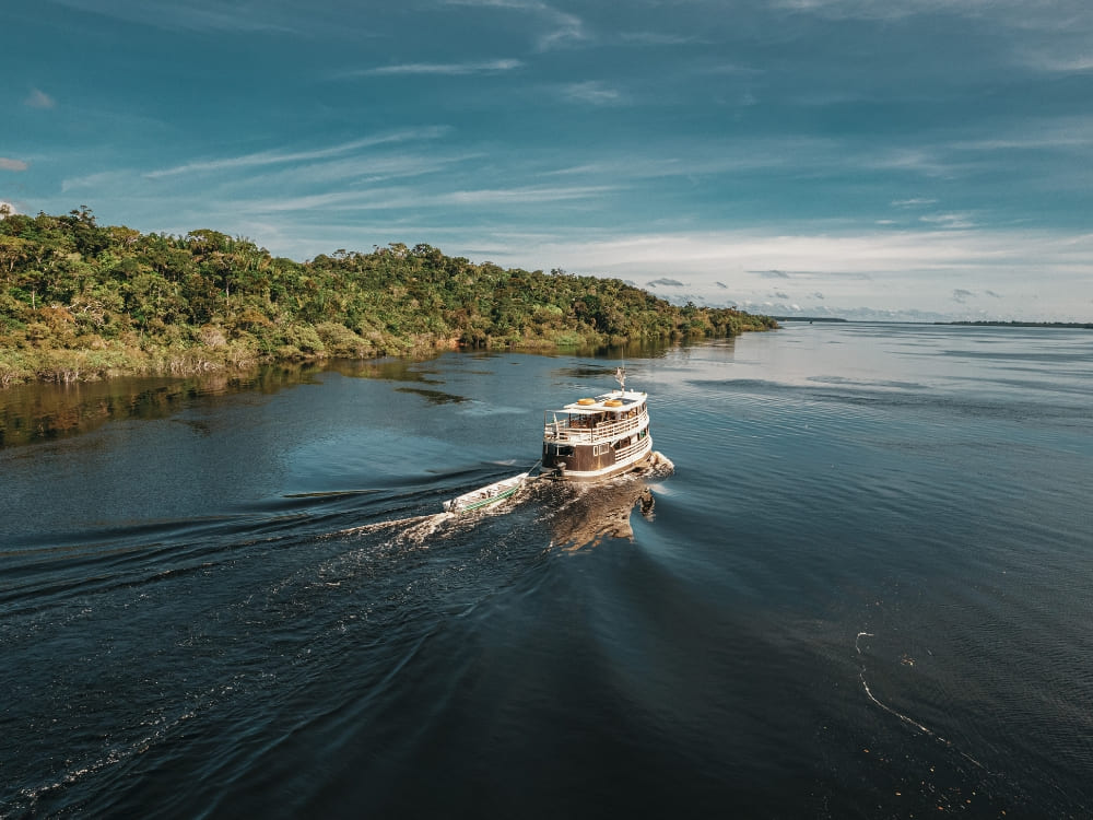 A river cruise boat navigating the calm waters of the Amazon River surrounded by lush green jungle. A river cruise boat navigating the calm waters of the Amazon River surrounded by lush green jungle.