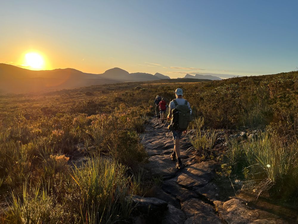 A group of hikers with backpacks walks along a rocky trail towards the mountains during a bright sunset in Chapada Diamantina. A group of hikers with backpacks walks along a rocky trail towards the mountains during a bright sunset in Chapada Diamantina.