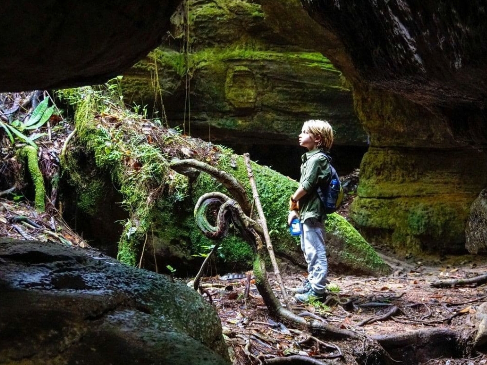 Young boy standing at the entrance of a mossy cave exploring the Amazon jungle.
