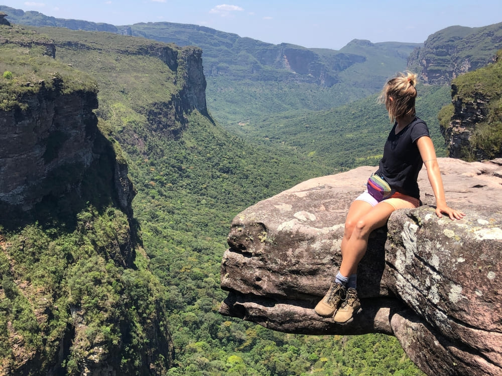 Woman hiker resting on a rocky cliff edge, overlooking a vast, lush green canyon and distant plateaus during an ecotourism adventure. Woman hiker resting on a rocky cliff edge, overlooking a vast, lush green canyon and distant plateaus during an ecotourism adventure.