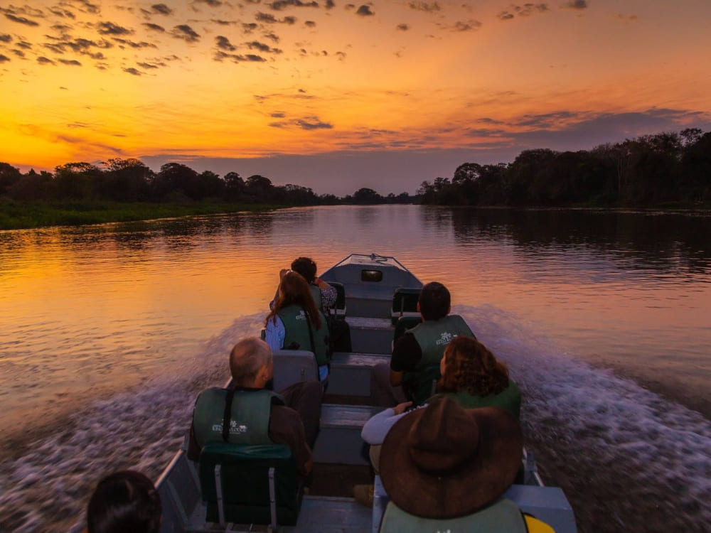 Travelers on a motorboat navigate a calm river during a vibrant orange sunset in the Pantanal