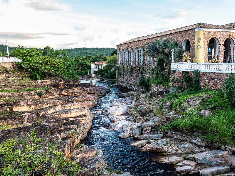 A river flowing over rocky layers next to a colonial building with stone arches in the historic town of Igatu.