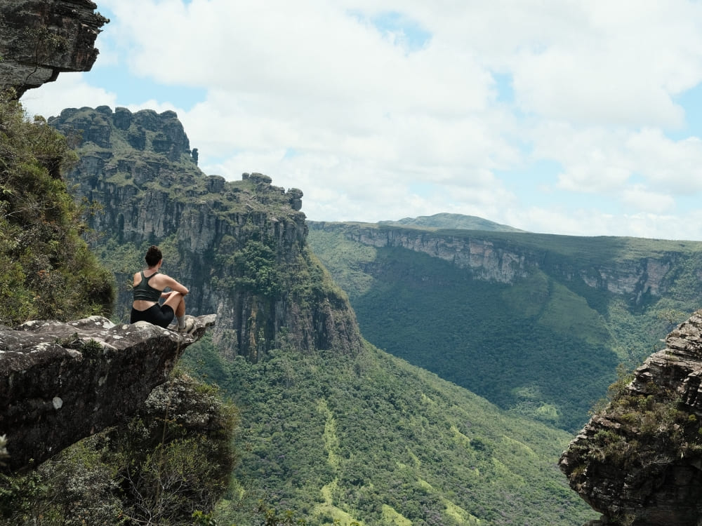 A person sits on a rocky cliff edge overlooking the deep, green forest canyons of Chapada Diamantina.