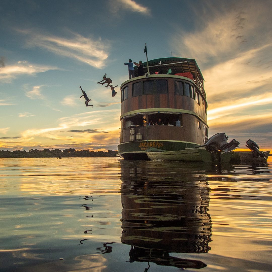 Tourists jumping into the river from the top deck of the Jacaré-Açu wooden boat at sunset during an authentic Amazon river cruise experience. Tourists jumping into the river from the top deck of the Jacaré-Açu wooden boat at sunset during an authentic Amazon river cruise experience.