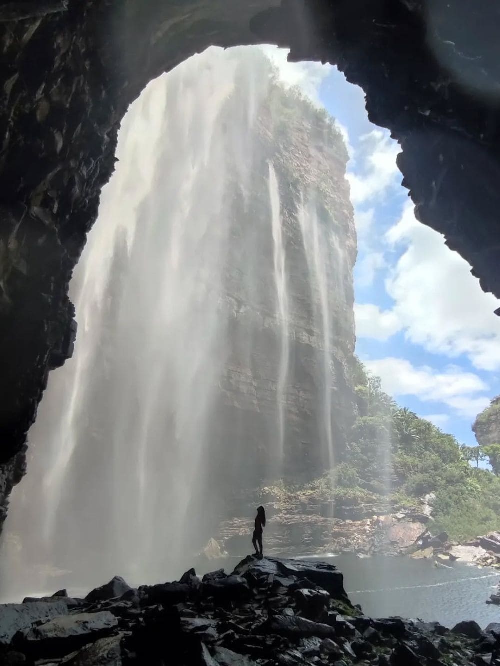 Silhouette of a hiker standing on rocks at the base of the massive Buracão waterfall flowing inside a canyon. 