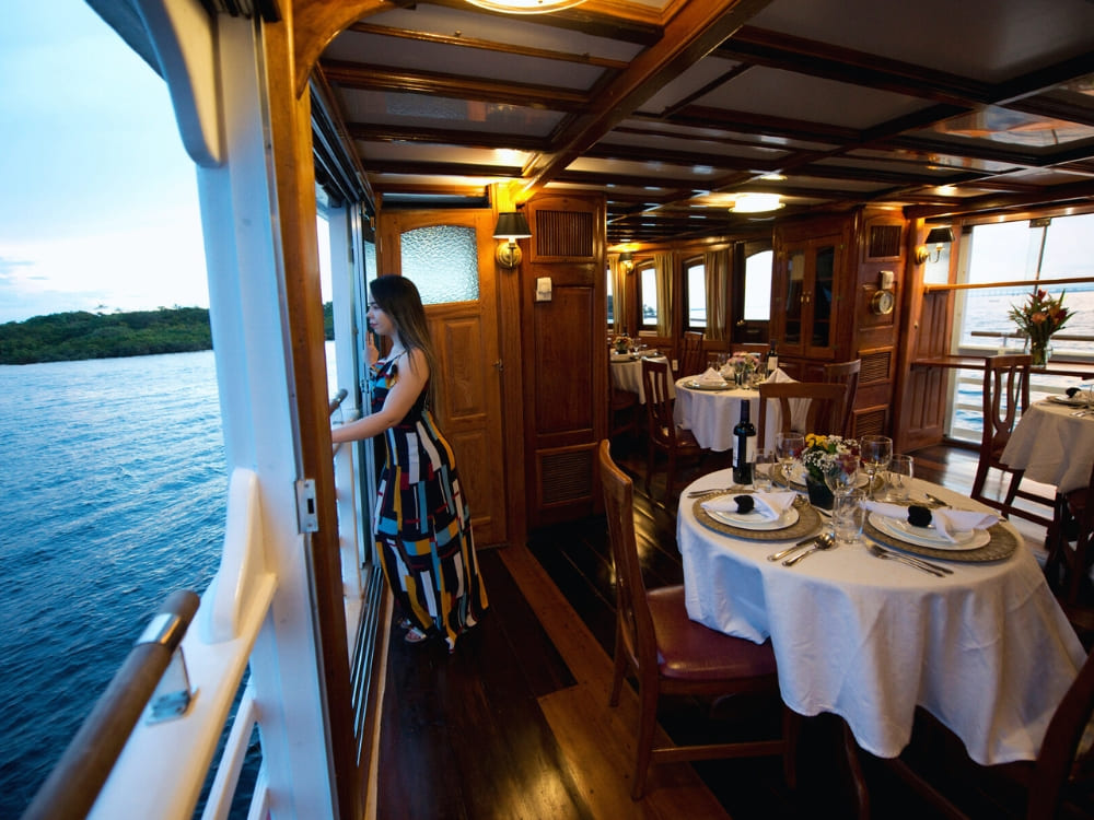 Breathtaking view of a colorful sunset over the Amazon River seen from the bow of the Tucano expedition boat. A woman in a colorful striped dress looks out the open window of a wooden boat dining room, which features tables set with white tablecloths and wine glasses.