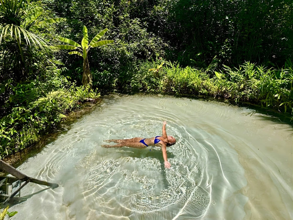 Woman floating on her back with her arms spread in crystal clear light blue waters, surrounded by tropical trees and dense green vegetation.