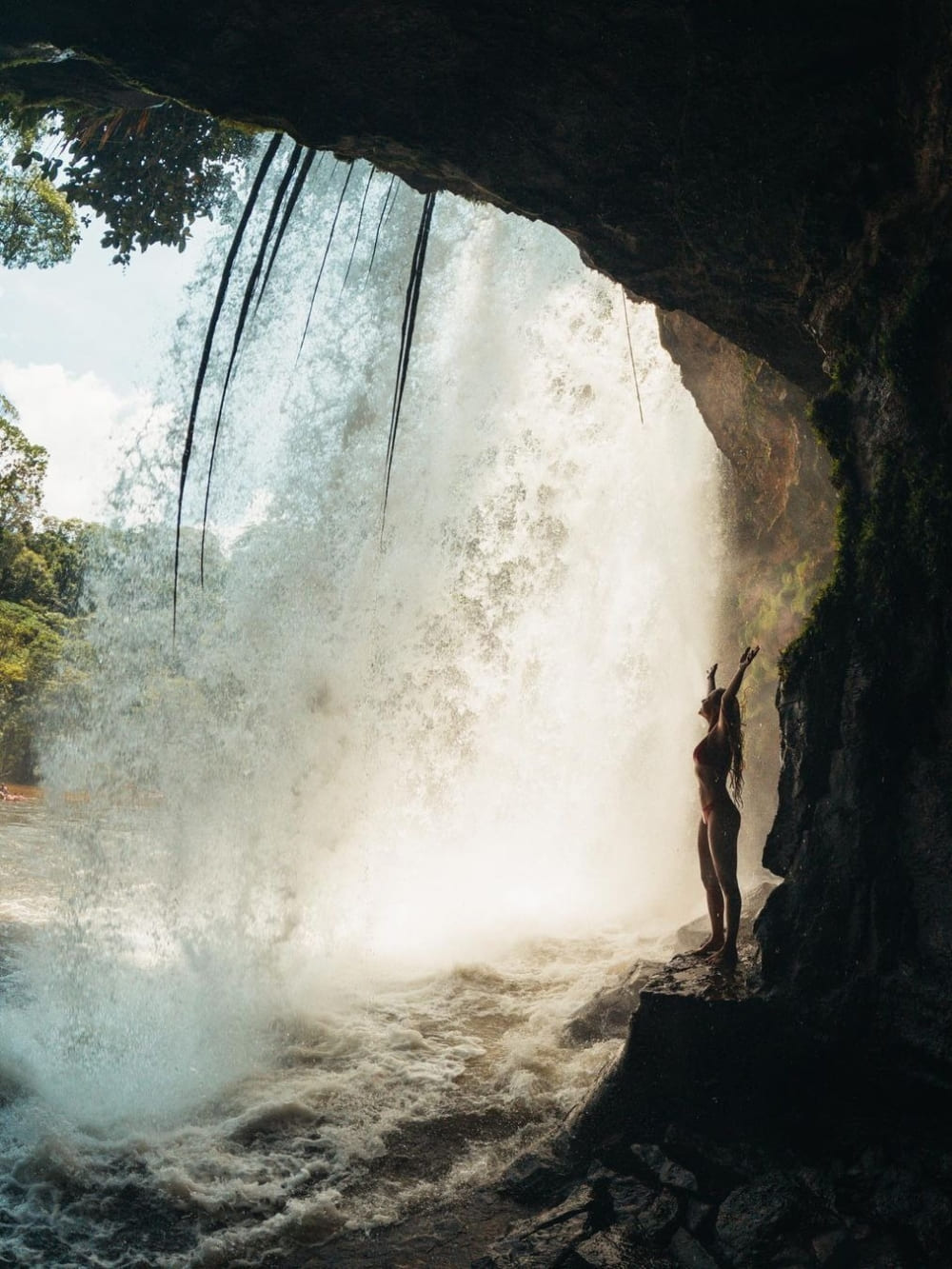 Silhouette of a woman with open arms contemplating a strong waterfall from the dark interior of a cave.
