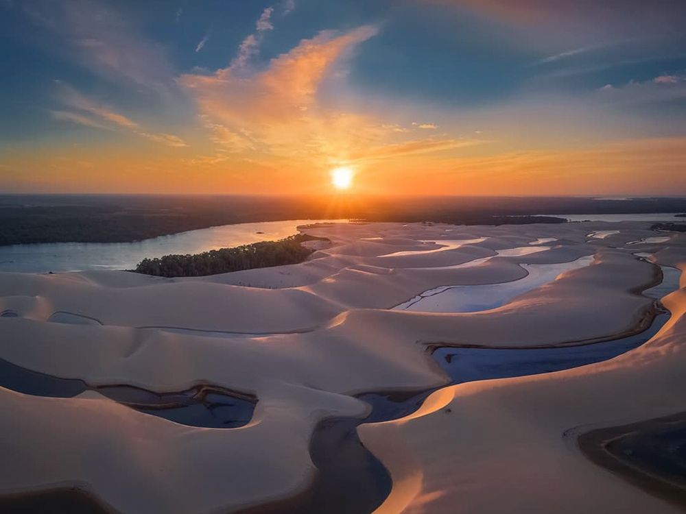 Aerial view of the dunes of Brazil with winding water channels reflecting the golden light of a vibrant sunset. 