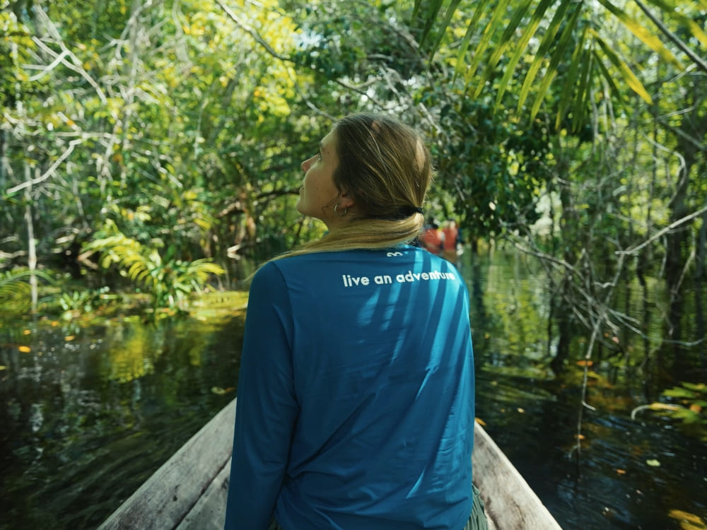  Woman from behind in a wooden boat navigating a river surrounded by dense forest, wearing a blue shirt with the phrase live an adventure.