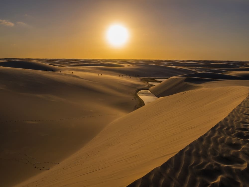 The sun sets brightly over undulating sand dunes and a shallow stream winding through the Lencois Maranhenses National Park in Brazil.