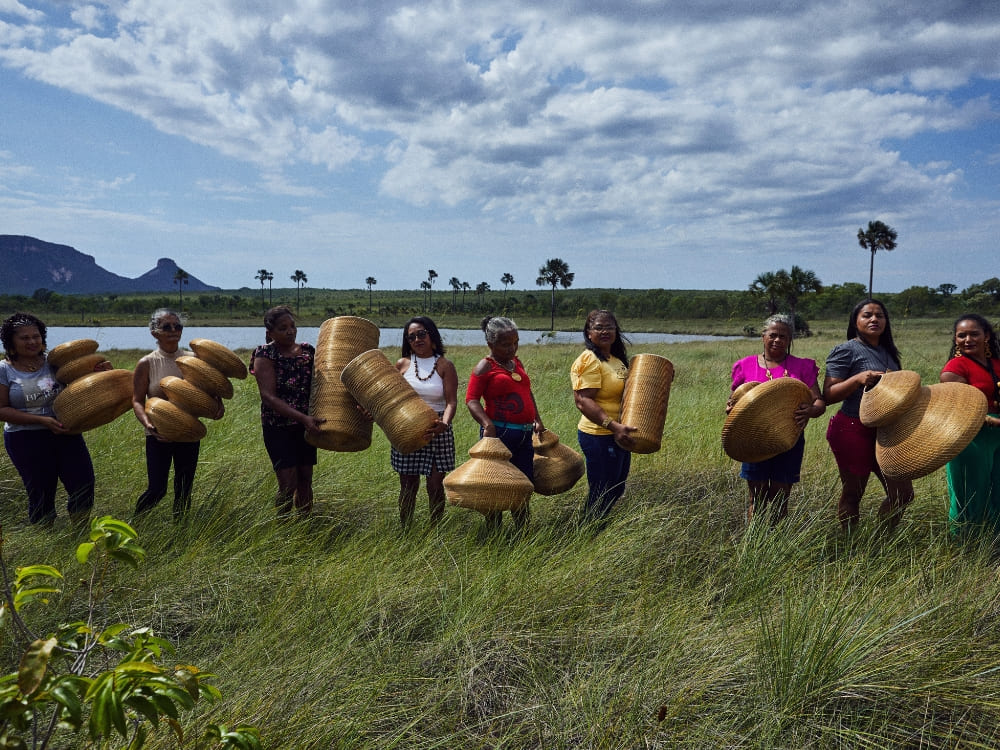 A group of women standing in a lush green field under a cloudy sky, holding up woven handicrafts made from Golden Grass in the Jalapão region. A group of women standing in a lush green field under a cloudy sky, holding up woven handicrafts made from Golden Grass in the Jalapão region.