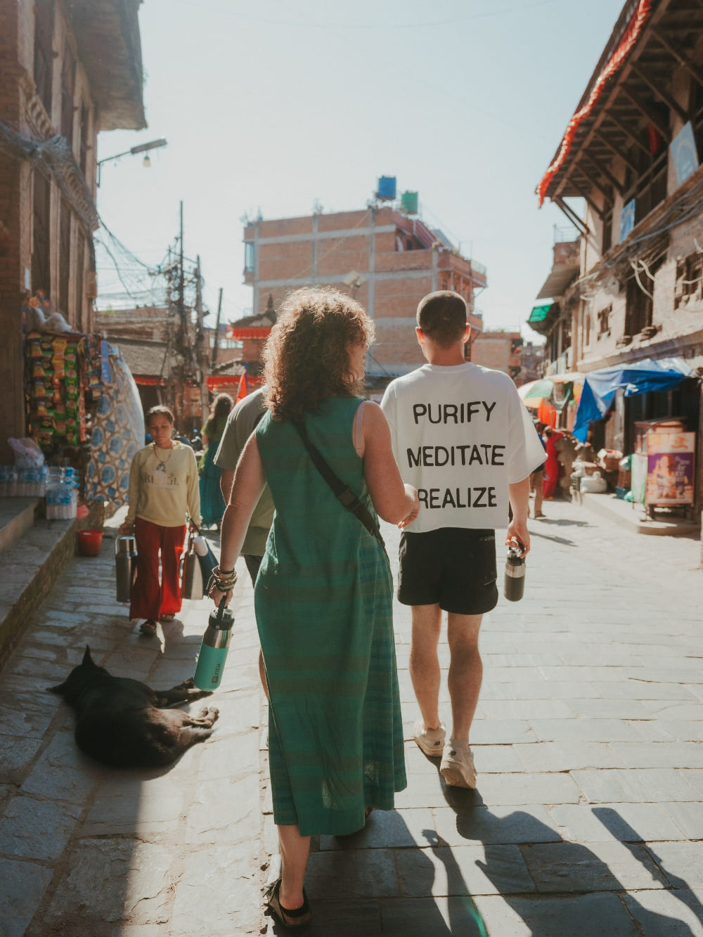 Mila and Lorenzo walking side by side down a sunny street in Nepal, with Lorenzo wearing a t-shirt that says purify, meditate, realize. Mila and Lorenzo walking side by side down a sunny street in Nepal, with Lorenzo wearing a t-shirt that says purify, meditate, realize.