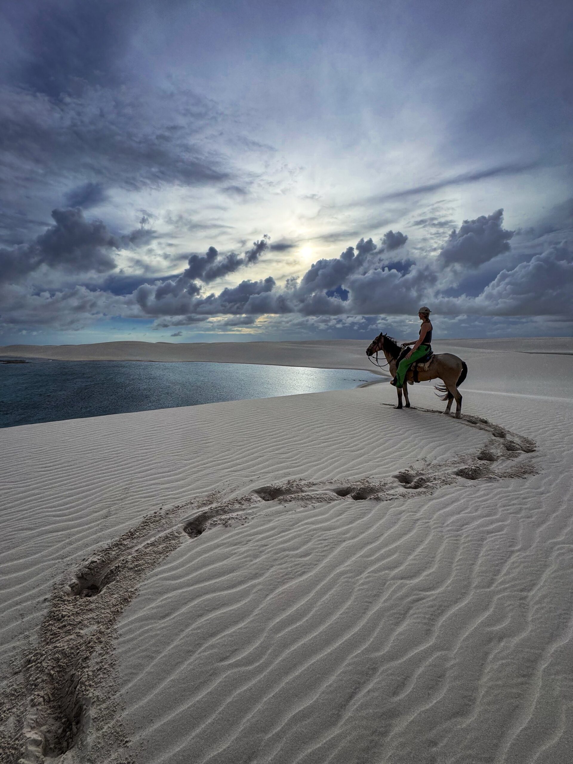 A solo rider crosses the white sand dunes of Lençóis Maranhenses National Park on horseback, with a rainwater lagoon stretching behind her.