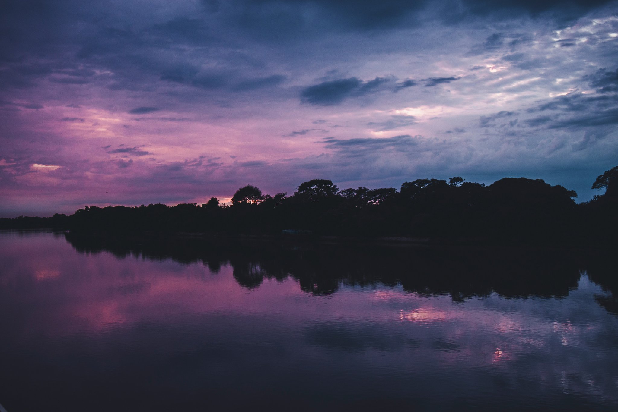 A vivid purple and pink sunset sky reflected on a calm river, with a dark treeline silhouette along the horizon on a pantanal tour from campo grande