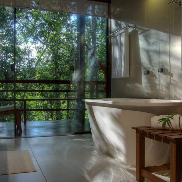 A freestanding bathtub beside a floor-to-ceiling window overlooking the rainforest in a luxury Amazon ecolodge bathroom.