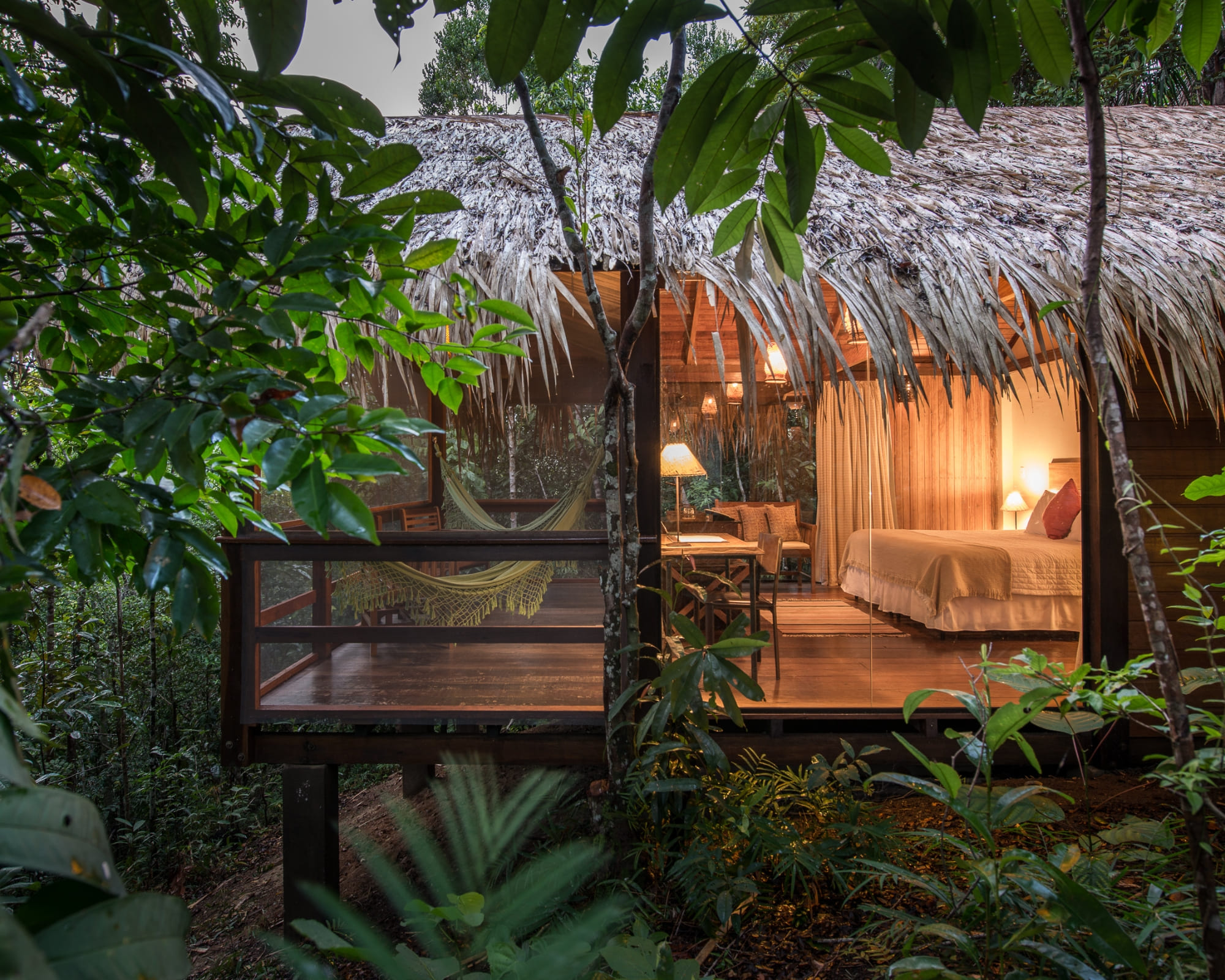 A luxury Amazon jungle lodge bungalow with a thatched roof and glass walls glowing with warm light at dusk, a hammock on its wooden deck amid the rainforest.