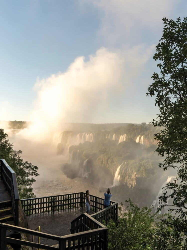 Dois visitantes na passarela do mirante sobre as Cataratas do Iguaçu ao amanhecer, no Brasil.