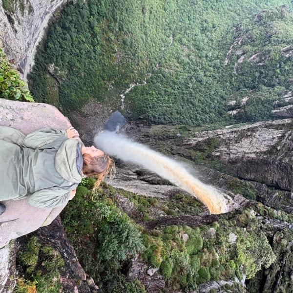 Trilheira deitada na rocha no alto da Cachoeira da Fumaça, espiando o abismo enquanto a água se desfaz em neblina sobre o vale verde, na Chapada Diamantina, Brasil.