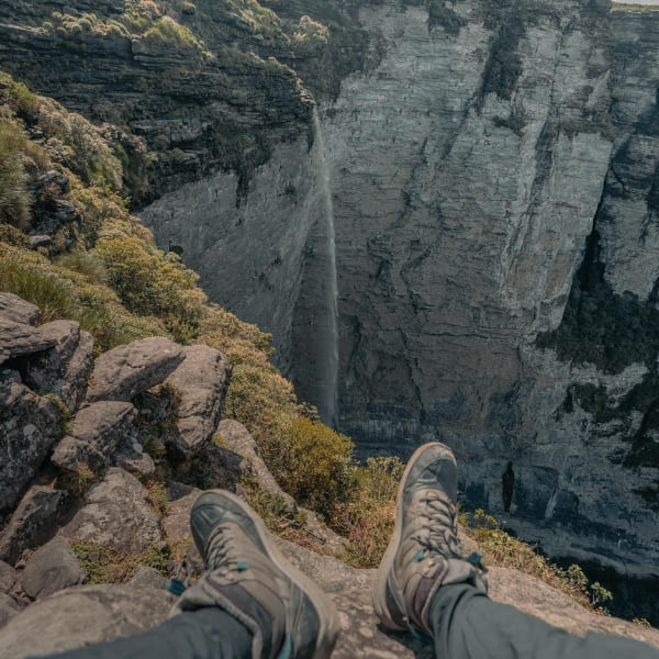 Botas de trilha na beira do penhasco no mirante da Cachoeira da Fumaça, com a queda d'água descendo pelo paredão do cânion em frente, na Chapada Diamantina, Brasil.