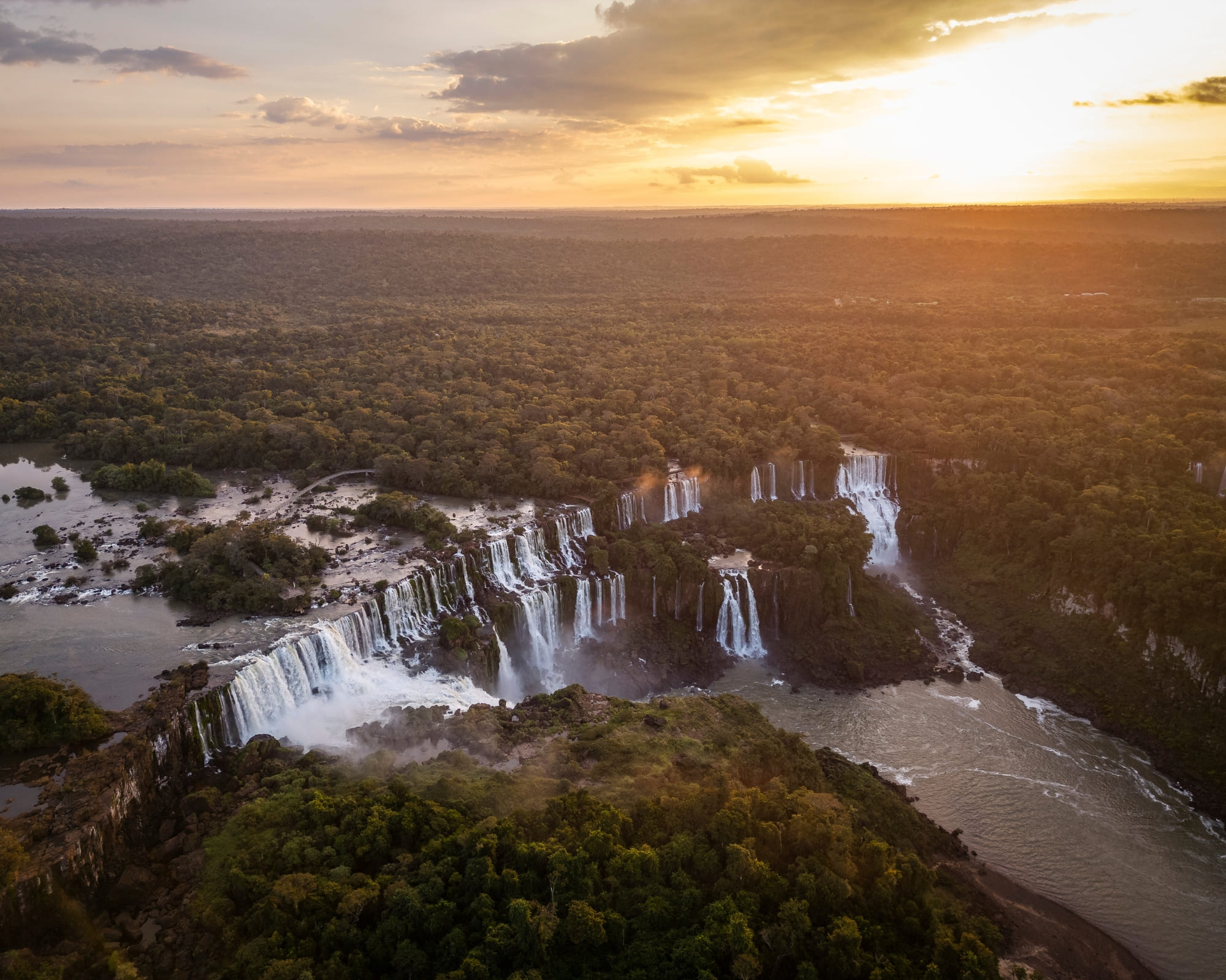 Vista aérea das Cataratas do Iguaçu ao pôr do sol, com dezenas de quedas cortando a Mata Atlântica, no Brasil.