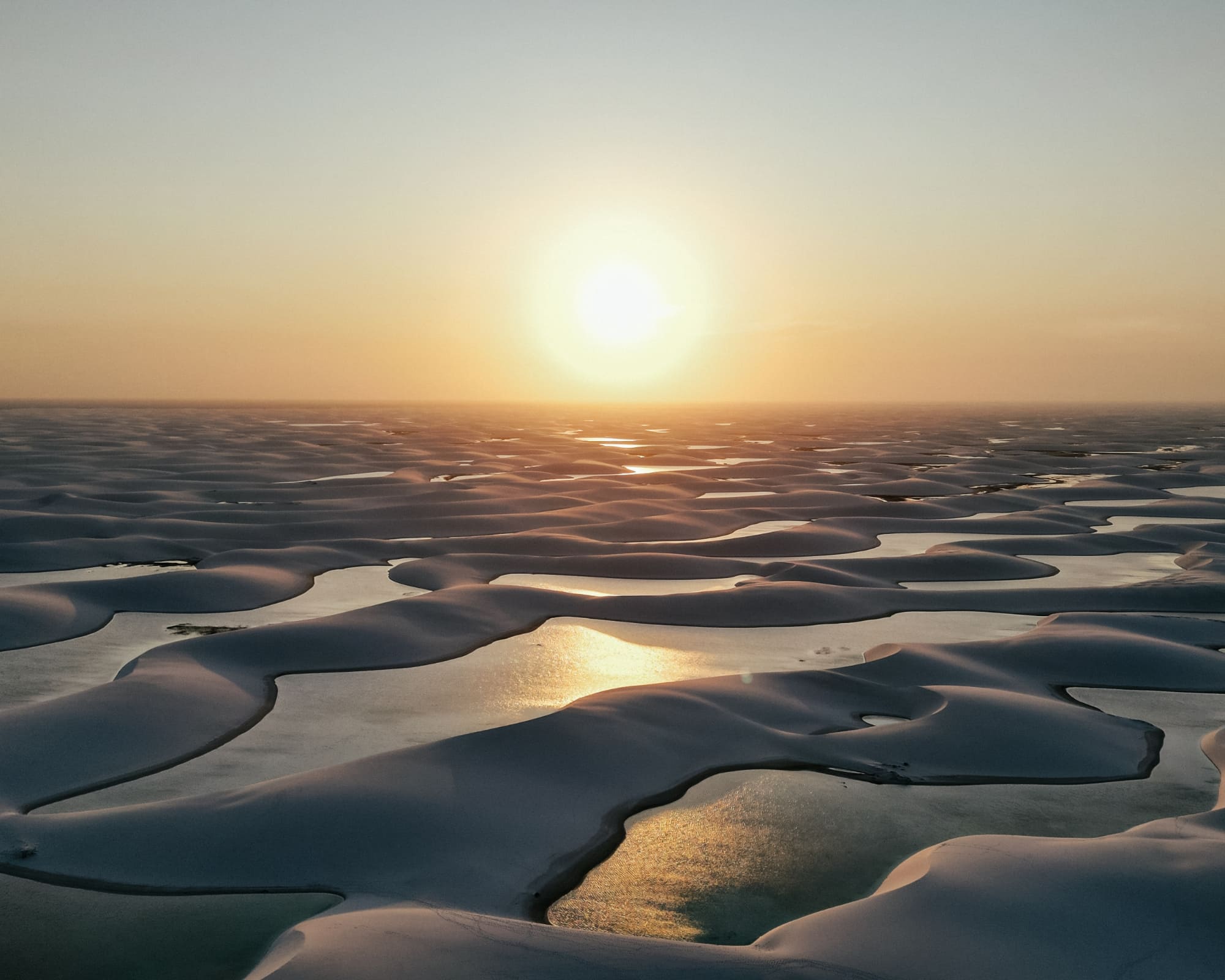 Vista aérea das dunas de areia branca e das lagoas de água de chuva dos Lençóis Maranhenses ao pôr do sol, Brasil.