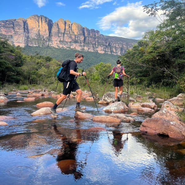 Dois trilheiros atravessando um rio raso sob os paredões da Chapada Diamantina, Brasil.