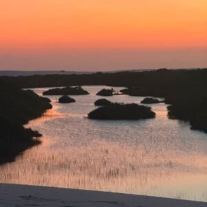 Cielo anaranjado reflejado en una laguna salpicada de islotes de vegetación, visto desde una duna en los Lençóis Maranhenses
